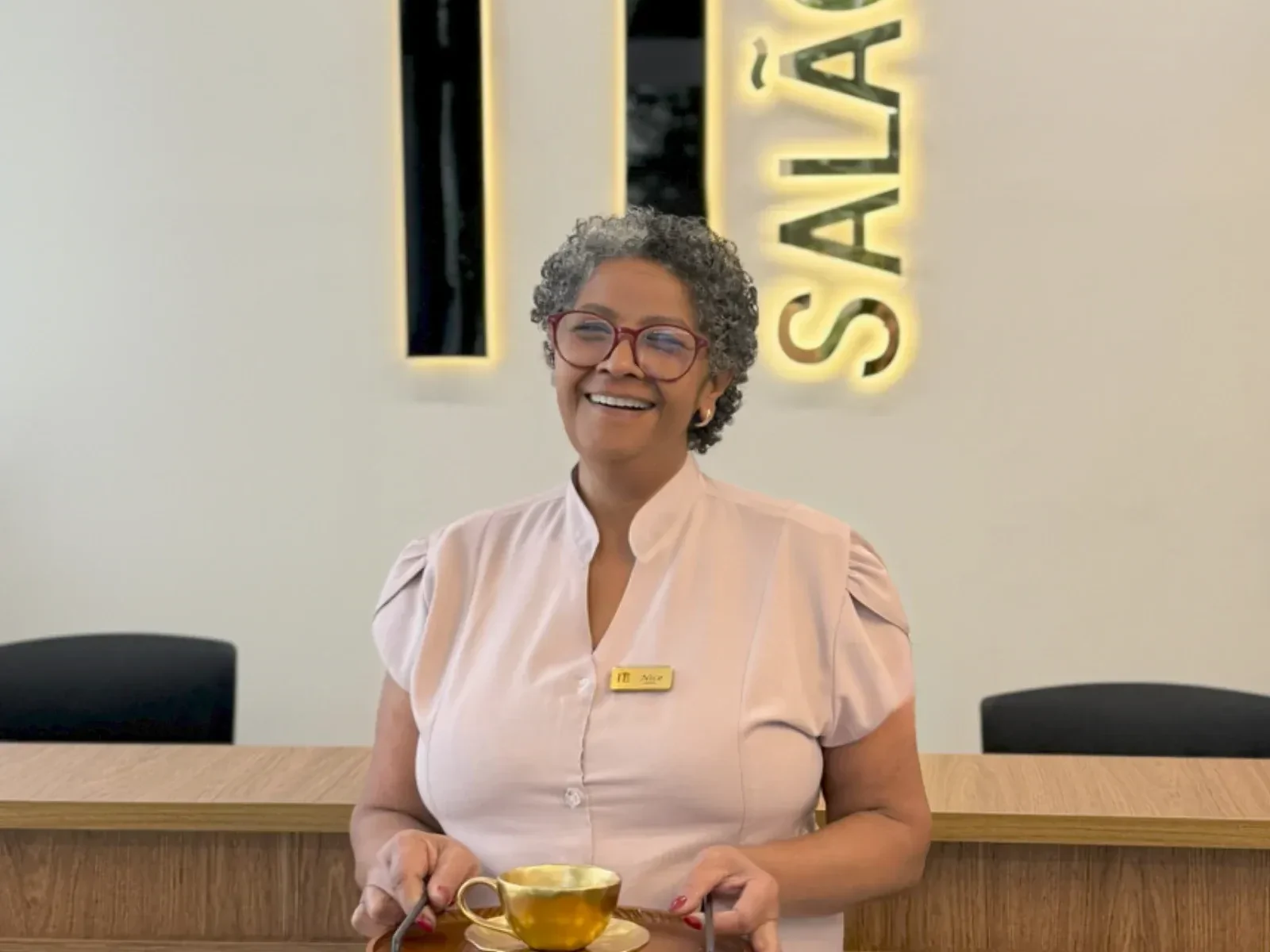 Recepcionista do IT Salão servindo café na recepção, sob o letreiro do salão
